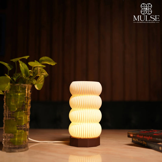 Modern ribbed 3D printed table lamp glowing on a wooden surface beside a small plant and books