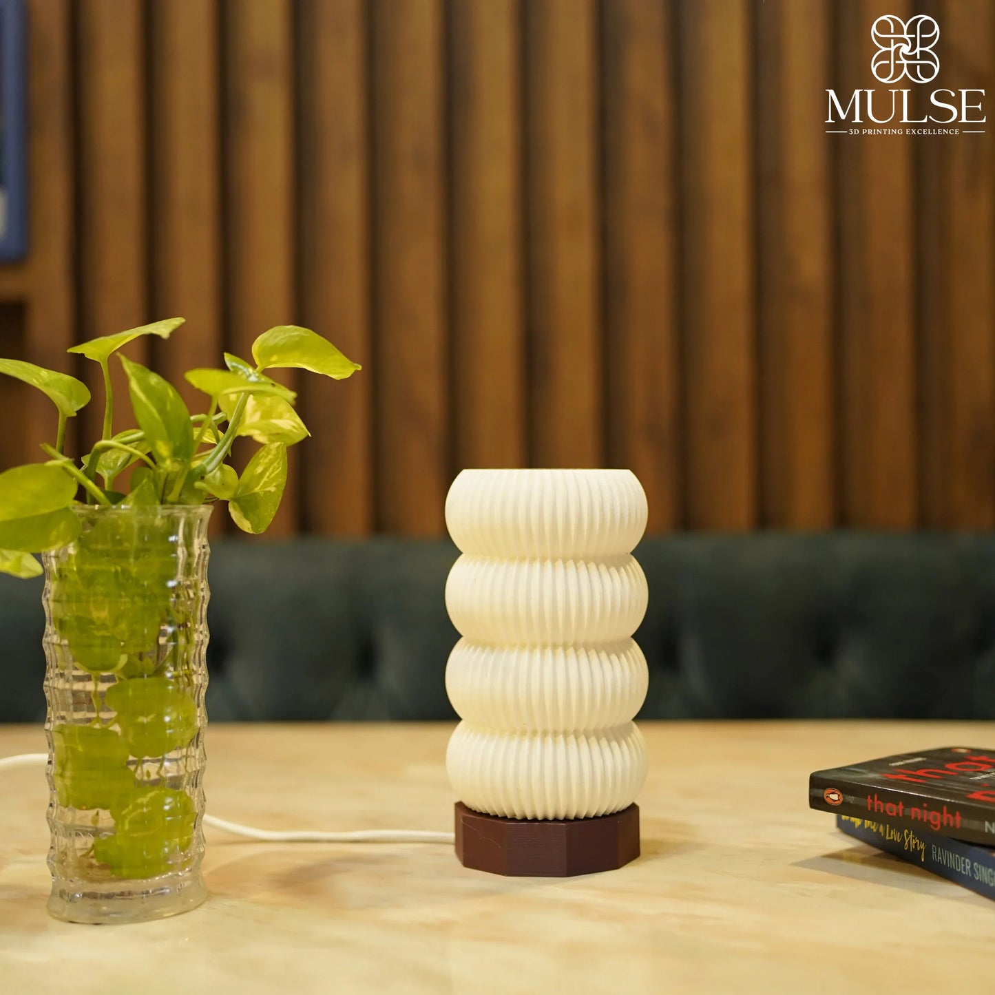 Modern 3D printed table lamp with ribbed texture next to glass vase with green plant on a wooden background